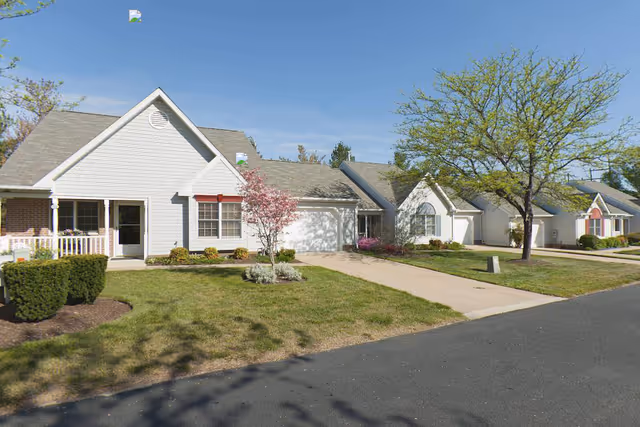 A row of single-story residential buildings with white siding and gray roofs, each with a driveway and small front yard featuring trees and shrubs. The sky is clear and blue.