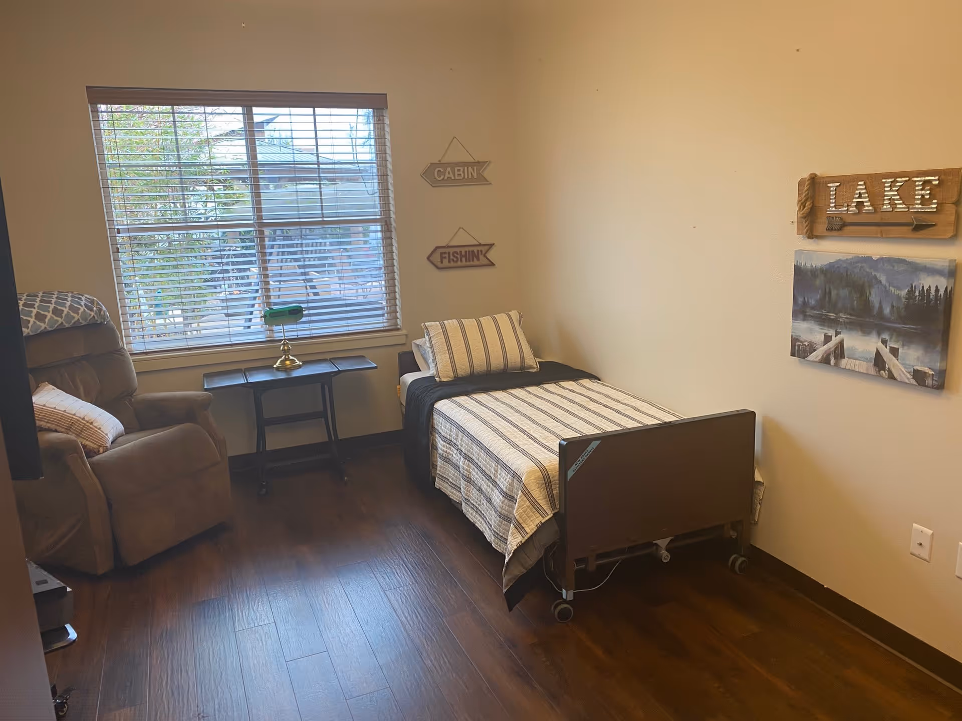 A small bedroom with a single bed covered in a striped bedspread and a matching pillow. Next to the bed is a brown recliner chair with a pillow. A window with blinds lets in natural light. On the wall above the bed are decorative signs that say 'CABIN', 'FISHIN'', and 'LAKE' along with a picture of a lake and dock. The floor is wooden.