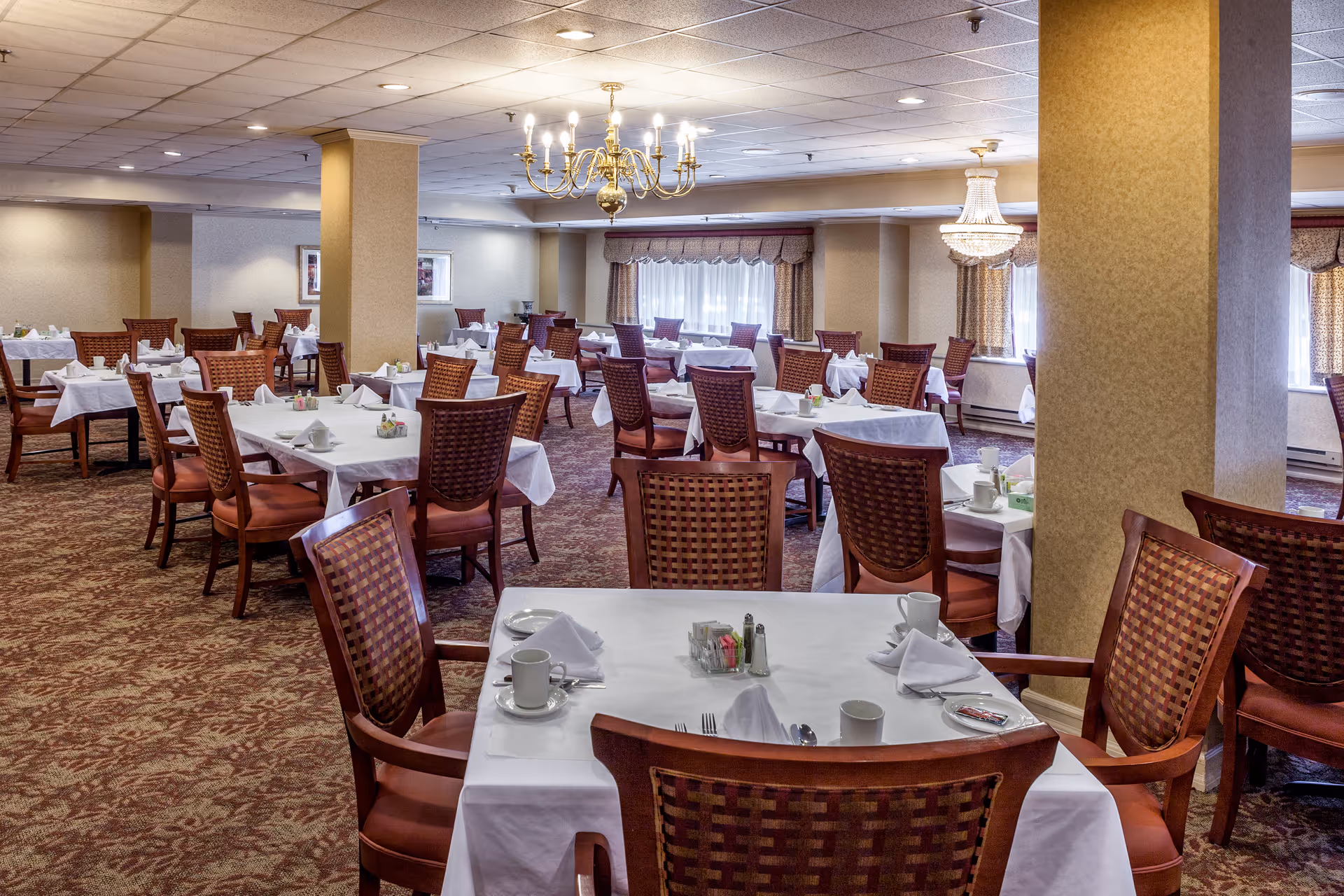 A spacious dining room in a senior living facility with multiple tables covered in white tablecloths, each set with cups, plates, napkins, and utensils. The room features patterned carpet, beige walls, large windows with curtains, and two chandeliers hanging from the ceiling. Wooden chairs with woven backs surround the tables.
