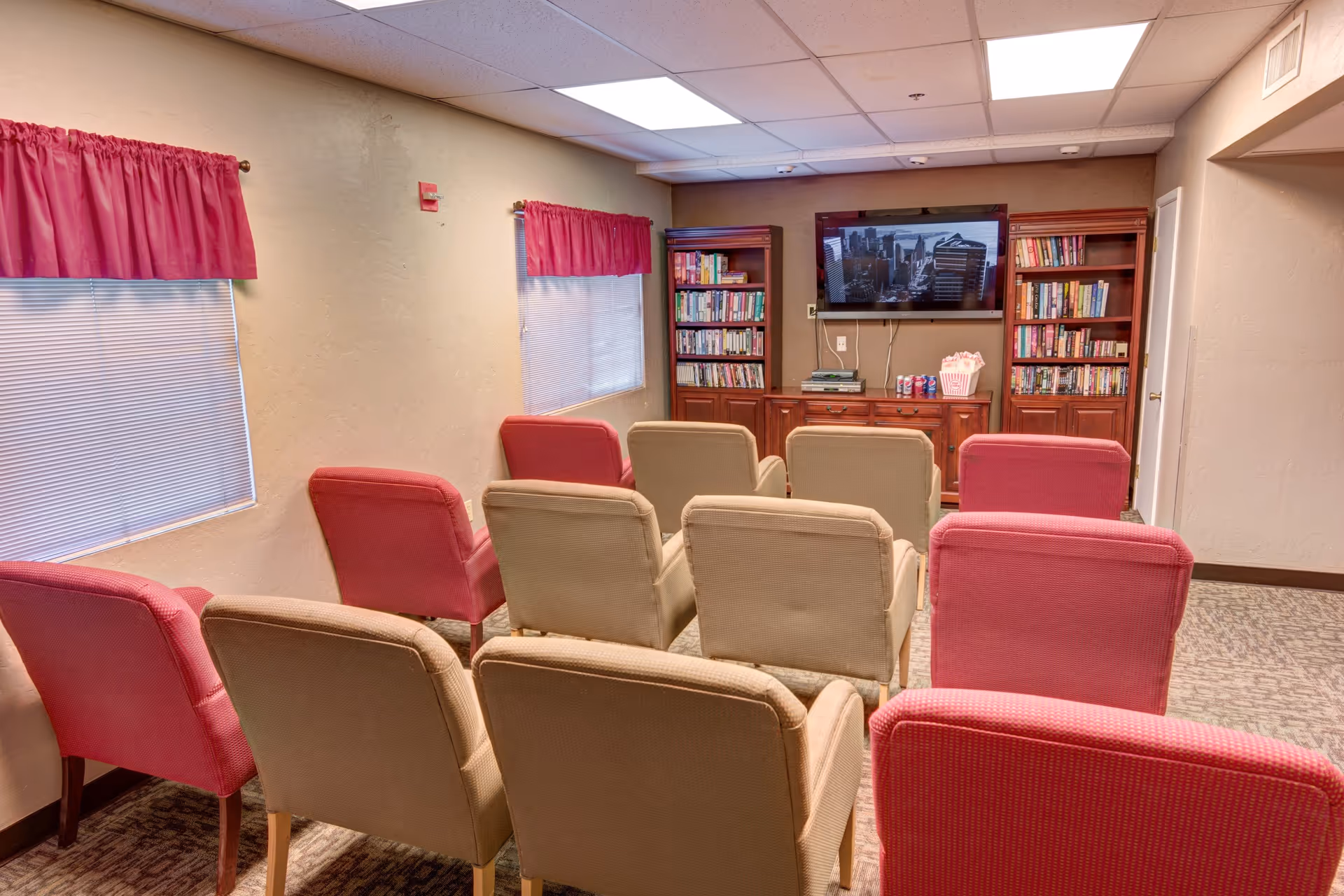 A small media room with two rows of beige and pink upholstered chairs facing a wall-mounted flat screen TV. On either side of the TV are wooden bookshelves filled with books. The room has beige walls, two windows with pink valance curtains, and a carpeted floor.