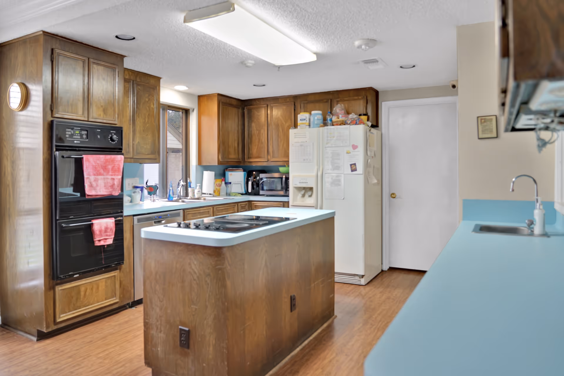 A kitchen with wooden cabinets, a white refrigerator covered with papers and magnets, a black double oven with red towels hanging on the handles, a kitchen island with a stovetop, and light blue countertops. The floor is wooden, and there is a window above the sink letting in natural light.