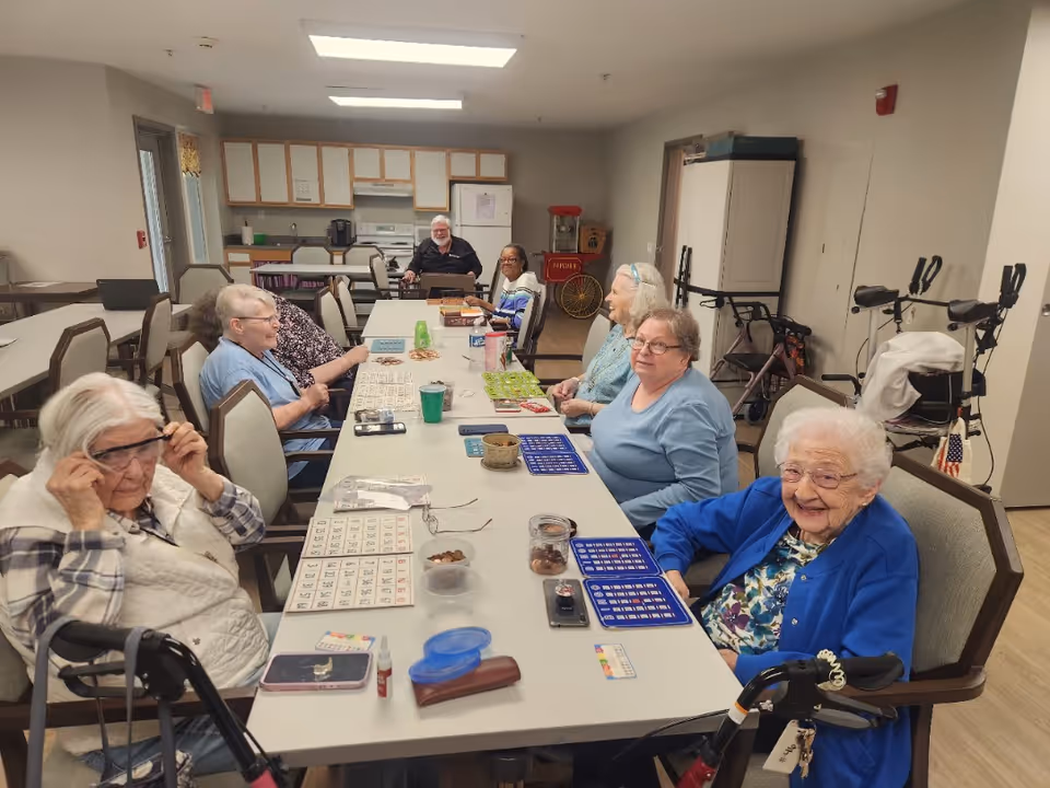 A group of elderly people sitting around a long table in a common room playing bingo. The room has a kitchenette in the background and mobility aids like walkers are visible. The atmosphere appears social and engaging.