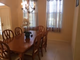 Sunlit dining room with a wooden table and six chairs, a chandelier overhead, a potted orchid centerpiece, and windows with sheer curtains.
