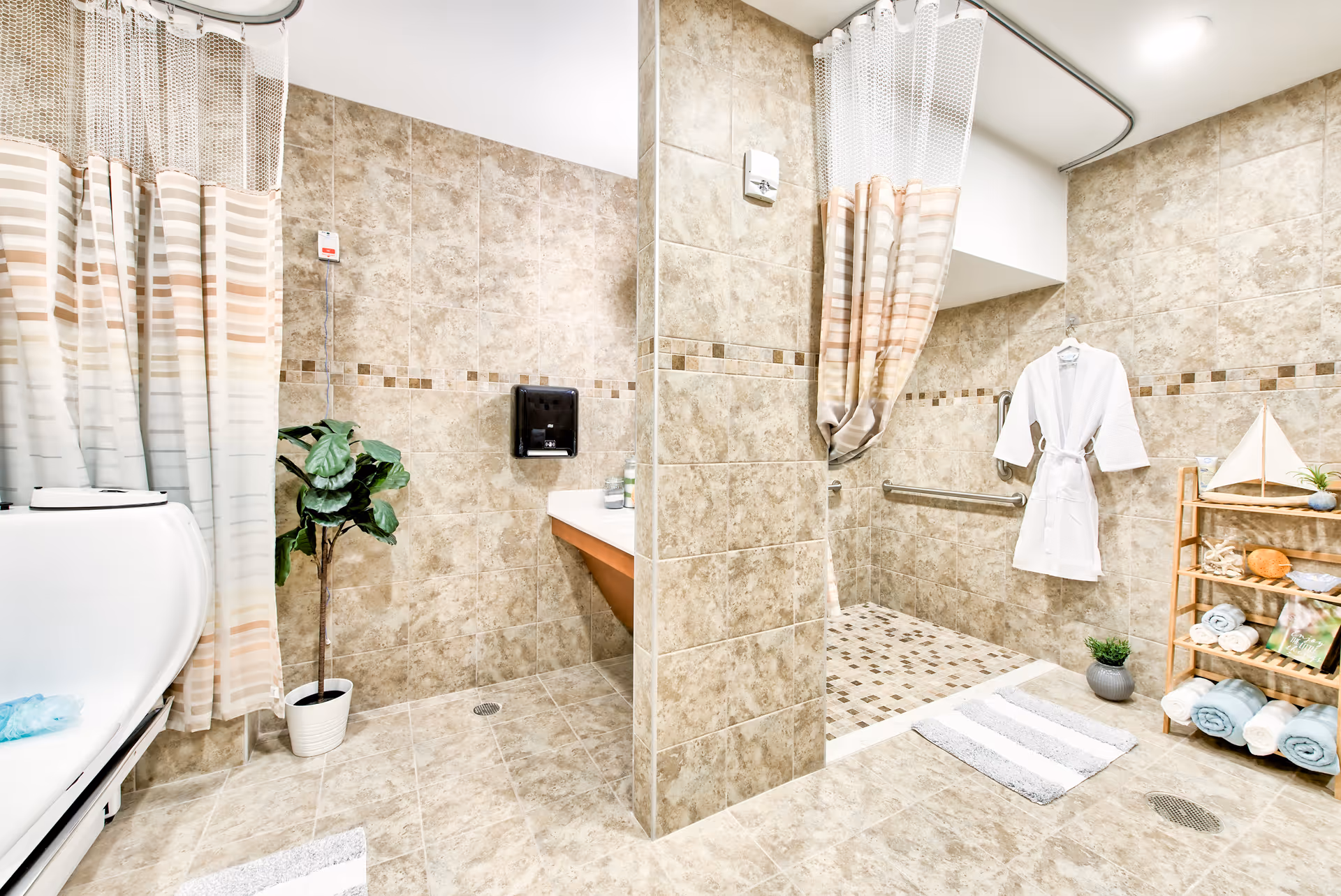 A spacious bathroom with beige tiled walls and floors, featuring a walk-in shower with a striped shower curtain, a white bathrobe hanging on the wall, a wooden shelf stocked with rolled towels and decorative items, a potted plant, and a white bathtub partially visible on the left side.