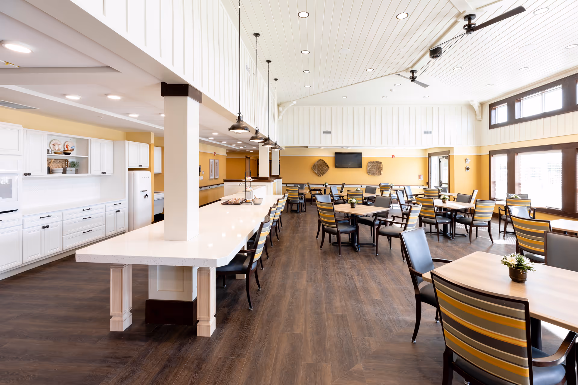 Bright and spacious dining area with multiple tables and chairs arranged neatly. The room features a long white countertop with bar stools, white cabinetry along one wall, large windows letting in natural light, and ceiling fans. The floor is dark wood, and the walls are painted a warm yellow with white paneling above.