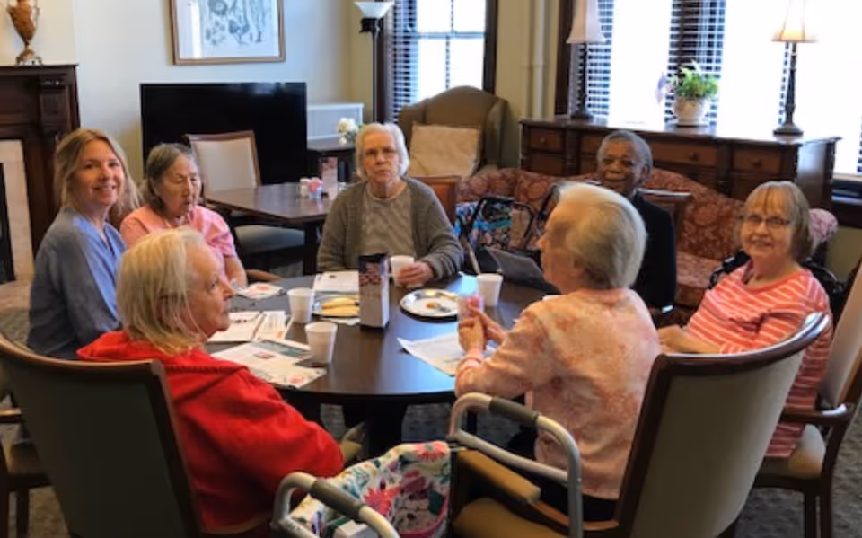 A group of elderly women sitting around a round table in a cozy living room or common area, engaging in conversation and activities with papers and cups on the table. The room has large windows with blinds, a TV, lamps, and comfortable chairs.