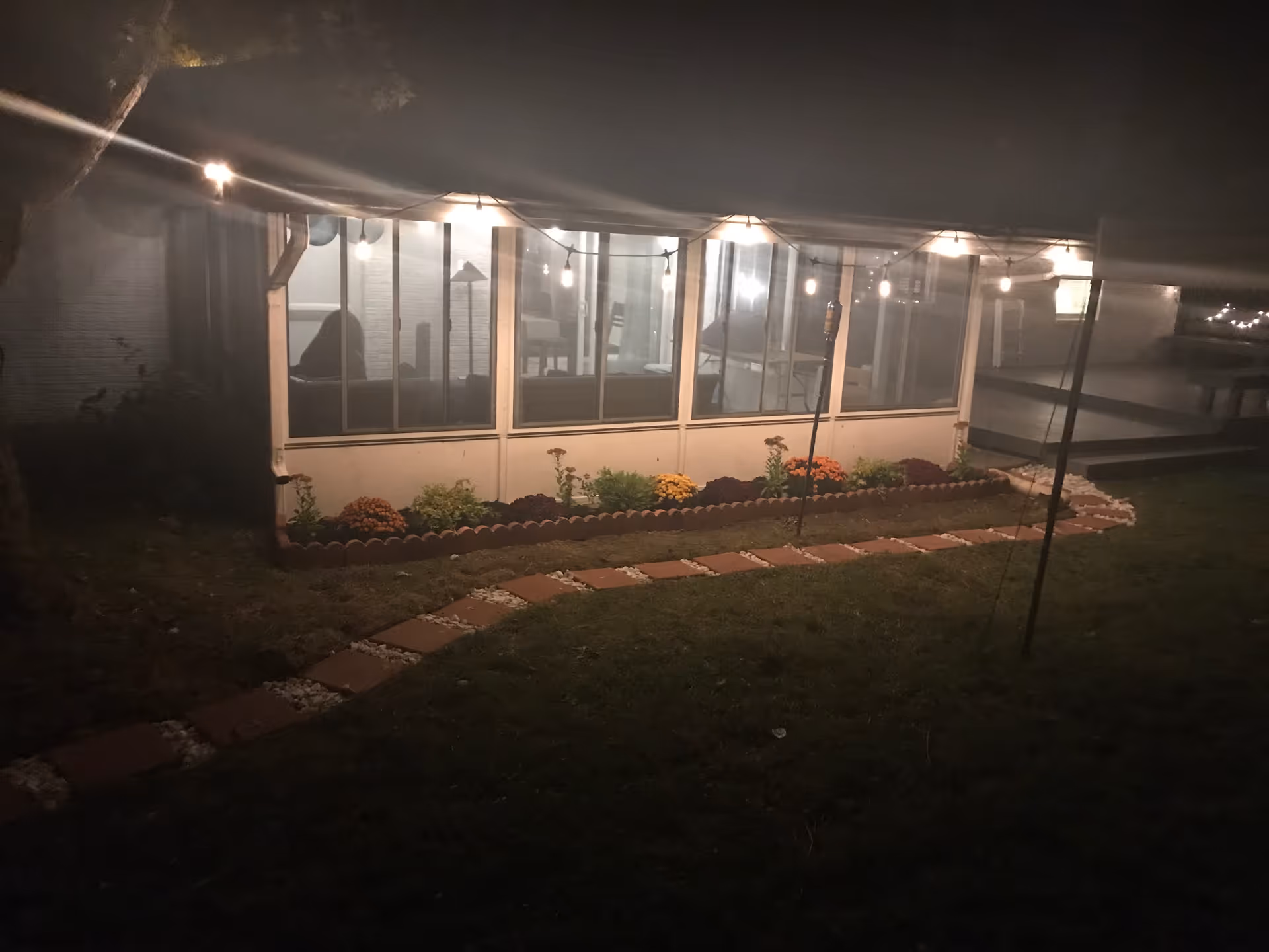 Lit enclosed porch/sunroom at night with string lights, a flower bed and a stepping-stone path across a lawn.