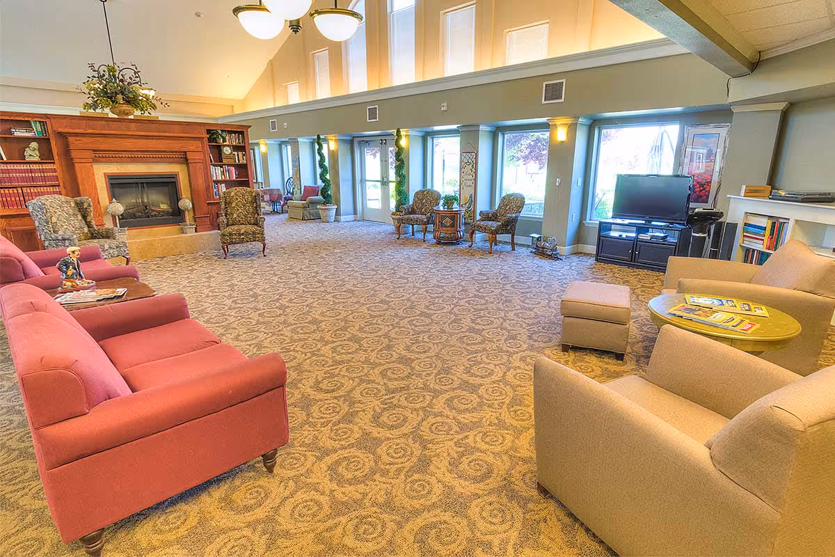 A spacious living room area in a senior living facility with patterned carpet, multiple upholstered chairs and sofas, a fireplace with built-in bookshelves, large windows letting in natural light, and a television on a stand.