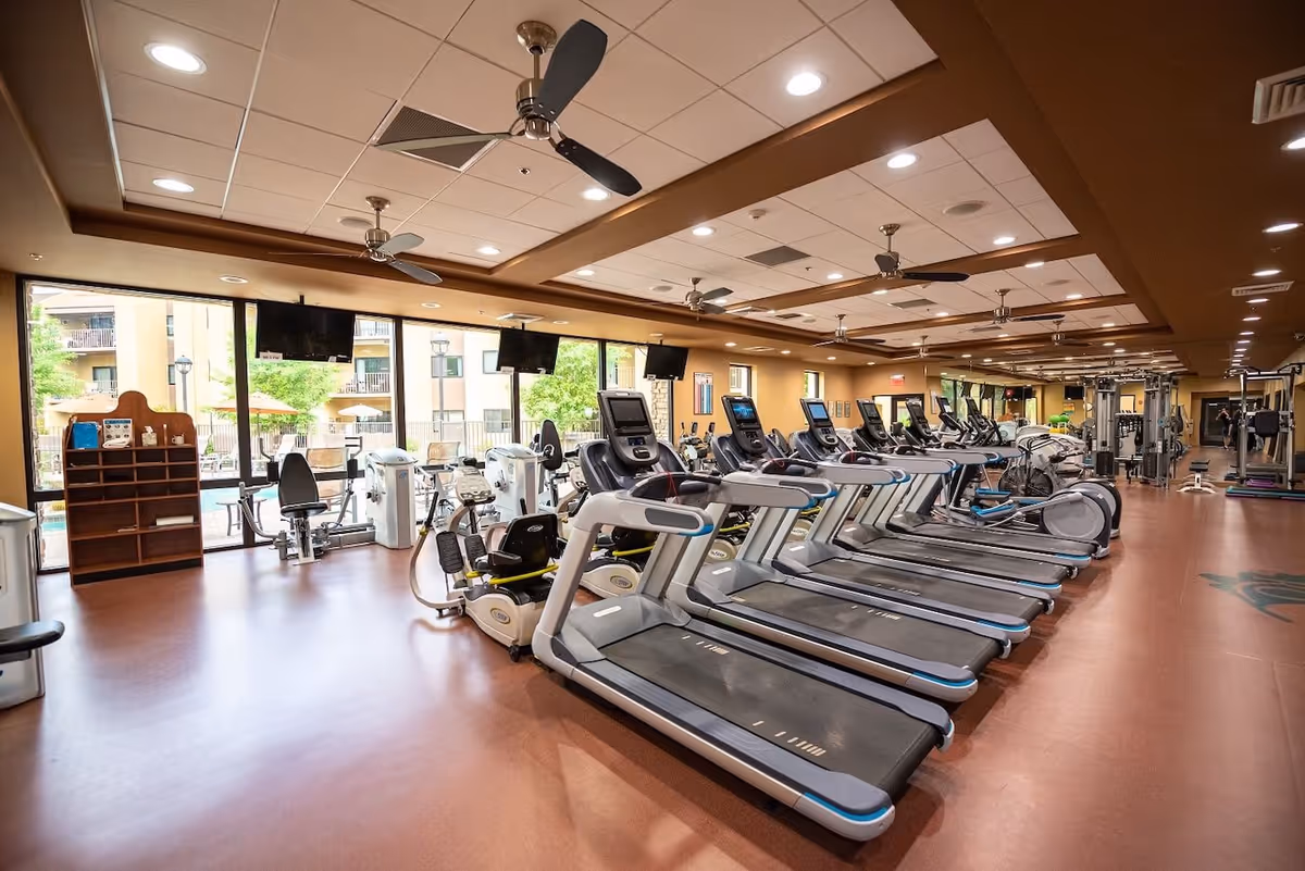 Spacious fitness center with rows of treadmills and other exercise machines under ceiling fans and large windows.