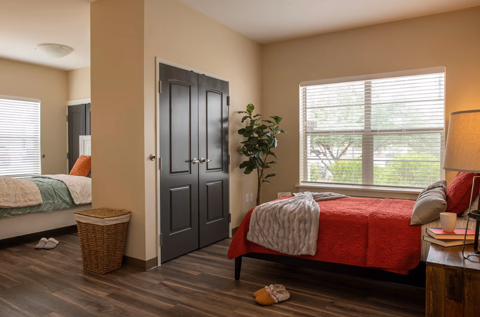 A cozy bedroom with a red quilted bedspread and a gray throw blanket on the bed. There is a wooden nightstand with a lamp, books, and a cup. A large window with blinds lets in natural light. A pair of slippers is on the floor near the bed. In the background, there is a second bed with a green bedspread and an orange pillow, partially visible through an open doorway. A wicker laundry basket is placed near the doorway, and a tall green plant stands next to a set of double dark-colored doors.