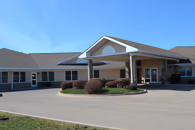 Exterior view of a single-story building with a covered entrance and a circular driveway. The building has beige walls, multiple windows, and neatly trimmed bushes in front. The sky is clear and blue.