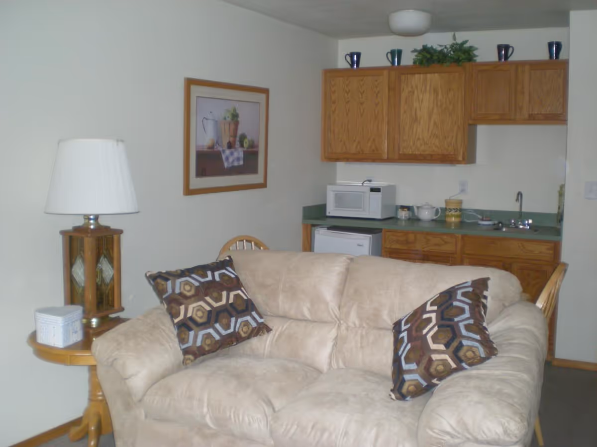 A cozy living area with a beige cushioned sofa adorned with two patterned pillows. Behind the sofa, there is a small kitchenette with wooden cabinets, a microwave, a mini fridge, and a sink. A wooden side table with a lamp and a small decorative box is placed next to the sofa. A framed picture hangs on the wall above the side table.