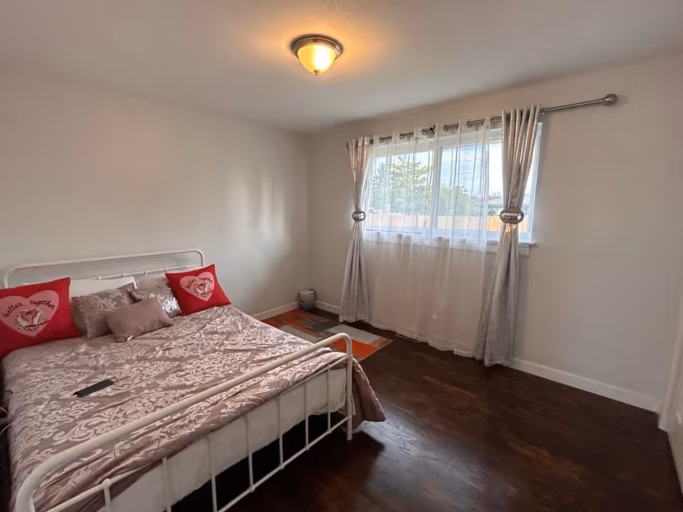 A bedroom with a white metal bed frame, a bed covered with a patterned beige comforter and several pillows including two red heart-shaped pillows with the words 'better together'. The room has dark wooden floors, a window with sheer white curtains and silver drapes, and a ceiling light fixture.