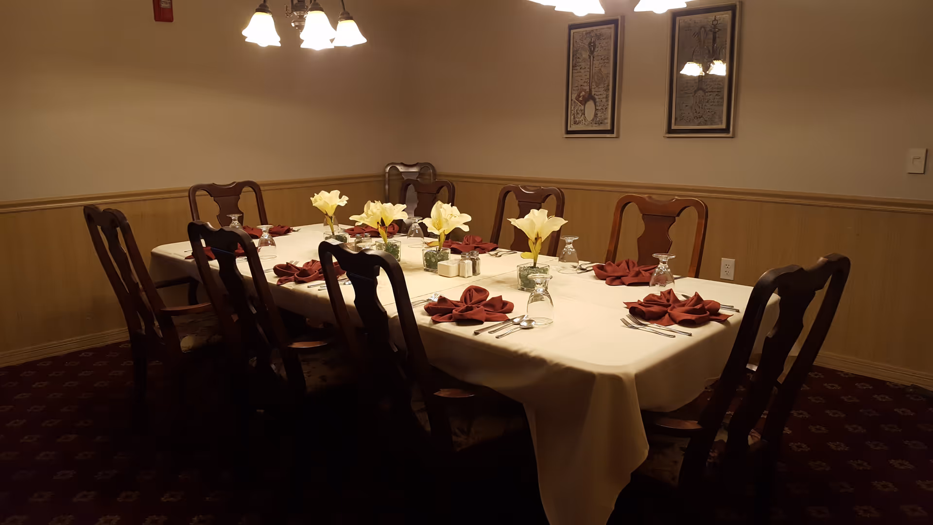 A formal dining room table set with place settings, folded napkins, and floral centerpieces under overhead lights.