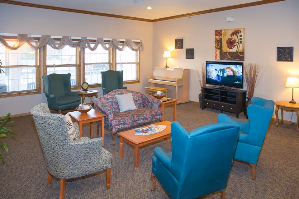 A cozy living room in an assisted living facility with multiple upholstered chairs and a floral-patterned loveseat arranged around wooden coffee and side tables. A television is on a stand against the wall, with a piano and decorative wall art nearby. Large windows with sheer curtains let in natural light, and lamps provide additional lighting.