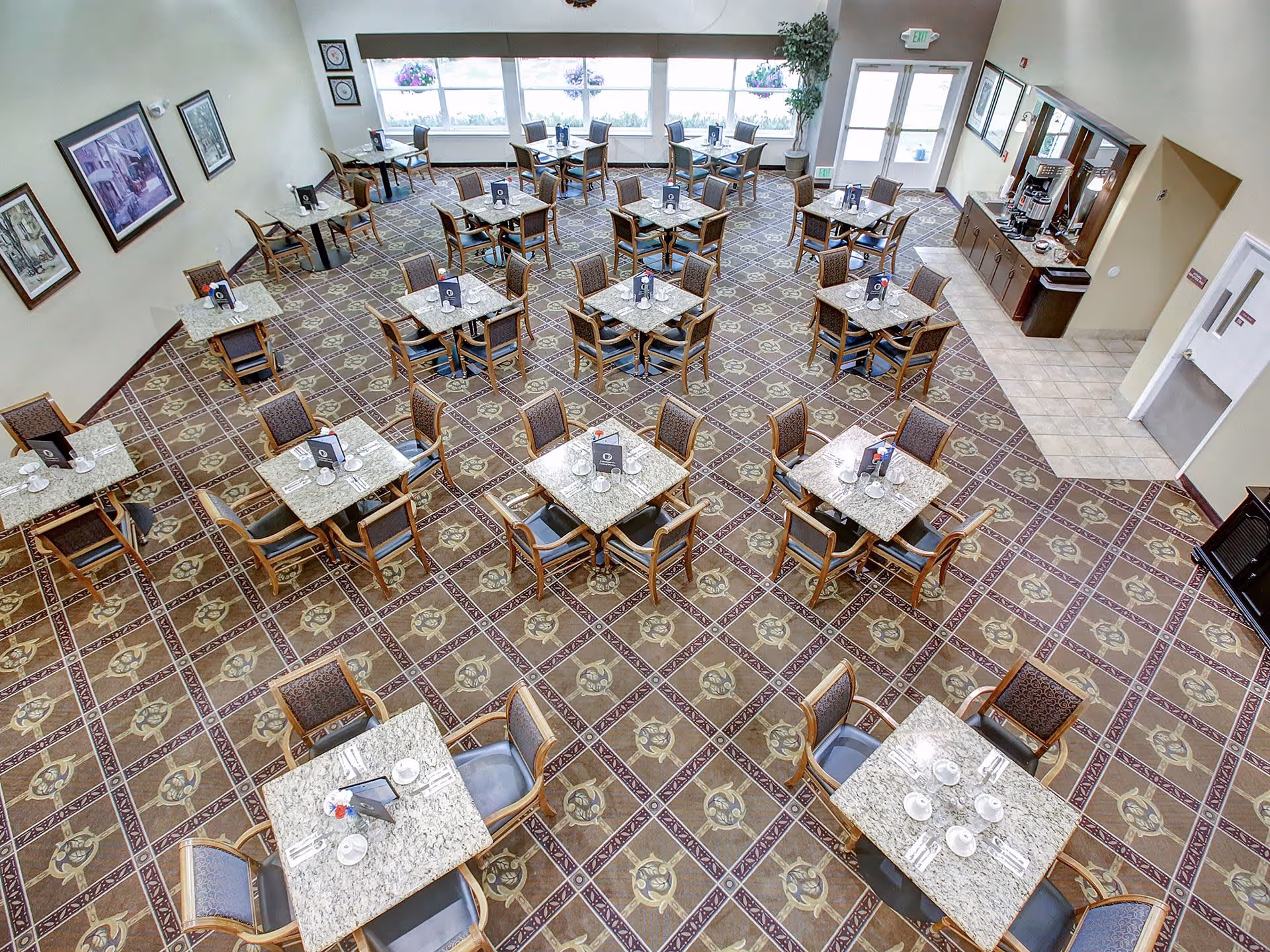 A spacious dining room with multiple square tables each set with cups, saucers, and utensils. The room has patterned tile flooring, large windows letting in natural light, framed artwork on the walls, and a beverage station with coffee machines and supplies near the entrance door.