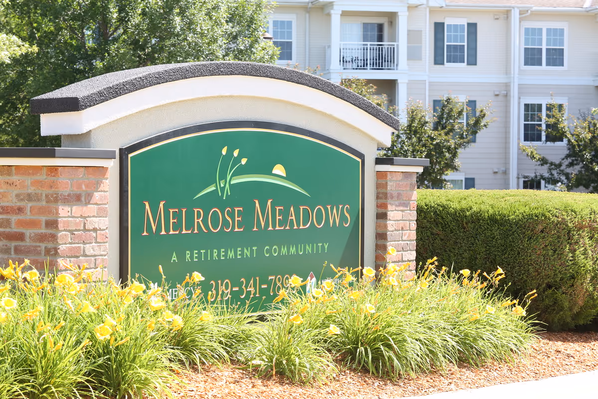 Entrance sign reading 'Melrose Meadows A Retirement Community' with yellow flowers and the building visible behind it.