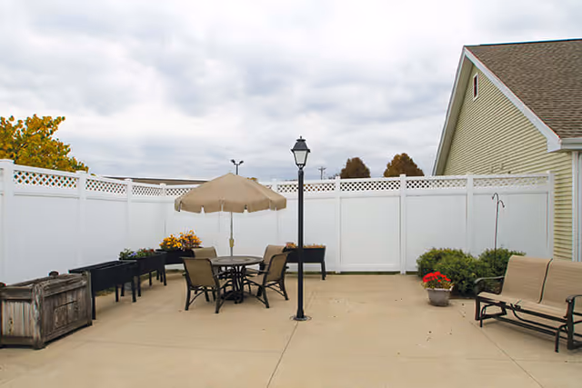 Outdoor patio area with a round table and four chairs under a beige umbrella, a black lamp post in the center, several planters with flowers and greenery, a wooden storage box, and a beige bench against a white fence and the side of a building.