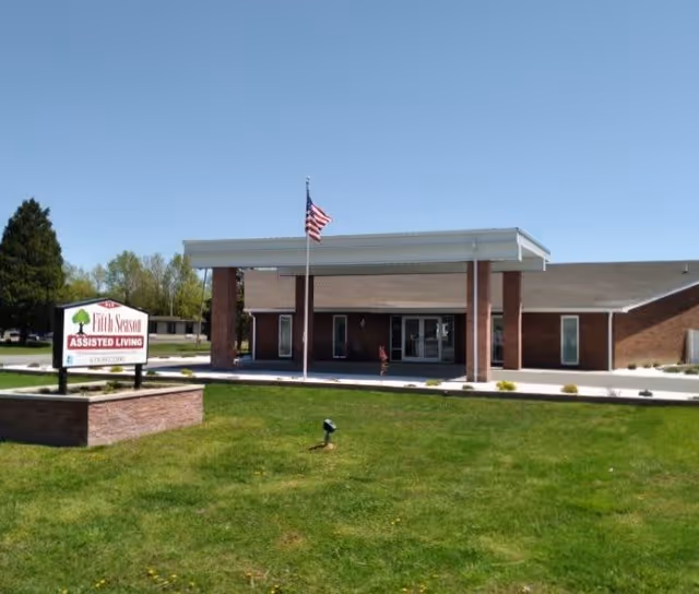 Exterior view of Fifth Season Assisted Living facility showing a single-story brick building with a covered entrance supported by brick columns. An American flag is flying on a flagpole in front of the entrance. There is a green lawn in the foreground and a sign on the left side that reads 'Fifth Season Assisted Living'.