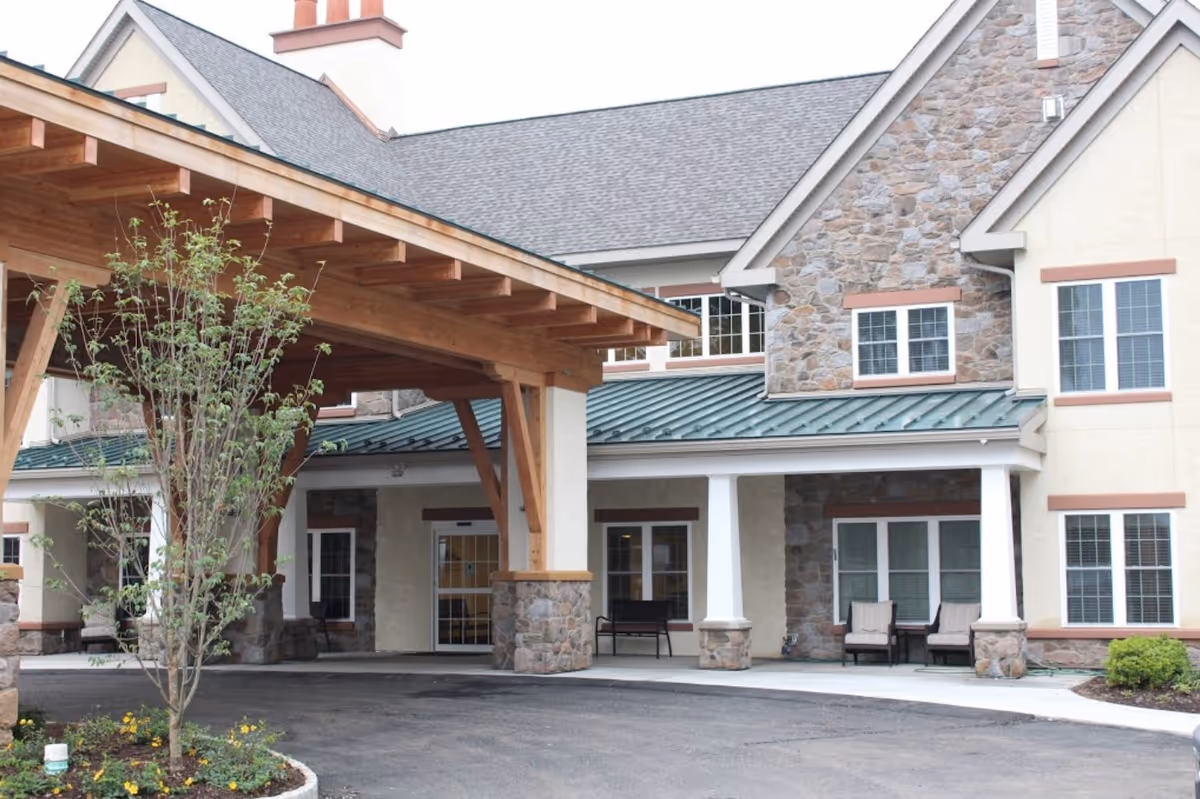 Entrance of Tel Hai Retirement Community featuring a covered driveway with wooden beams, stone pillars, and a landscaped area with a small tree and flowers. The building has a stone and beige exterior with multiple windows and outdoor seating.