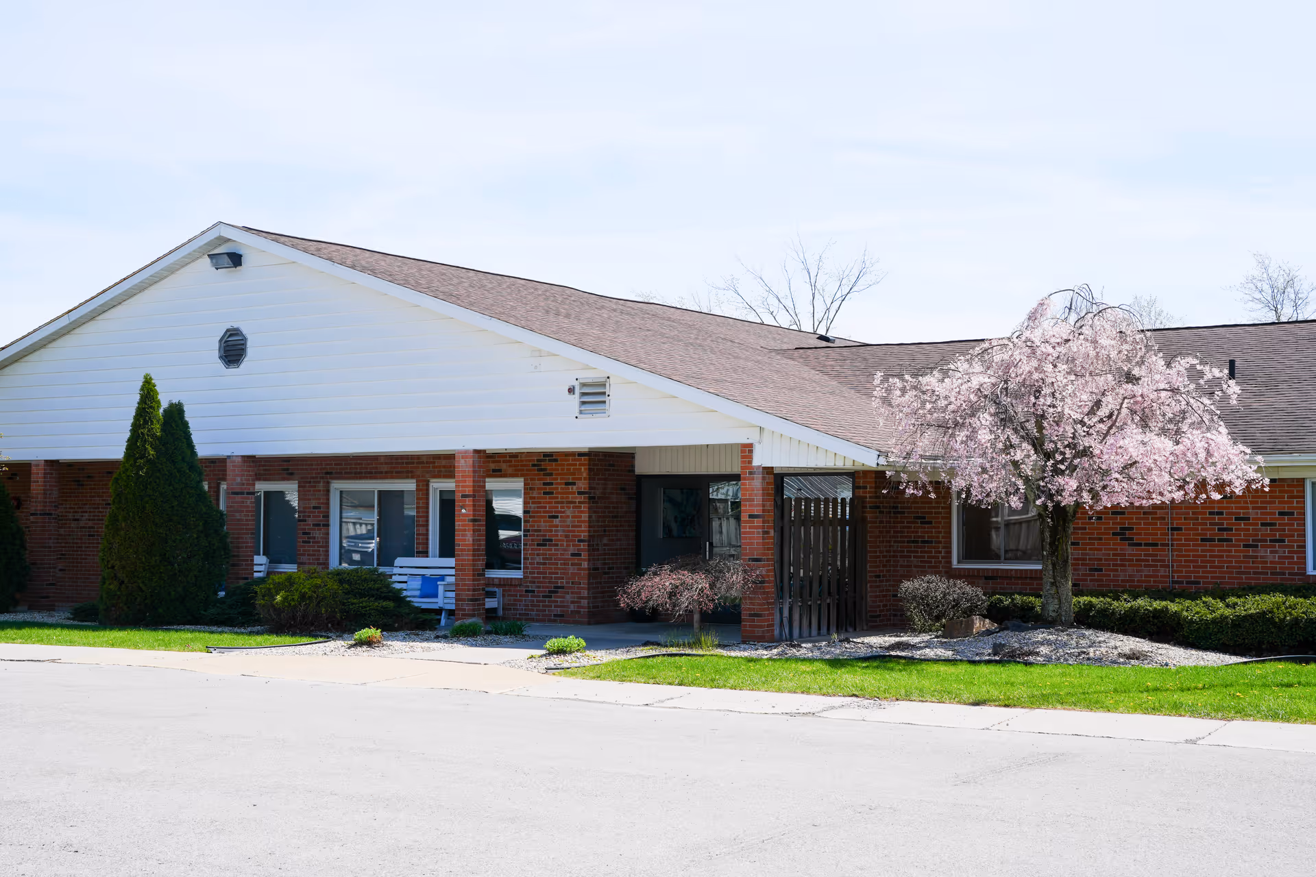 Single-story brick senior living facility entrance with a covered portico, landscaping and a blooming pink tree.