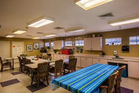 A well-lit dining room with multiple tables and chairs arranged for group seating. One table in the foreground is covered with a blue and green patterned tablecloth. The room has beige walls, several cabinets along one side, windows with blinds, and various decorations and plants on the countertops and shelves.