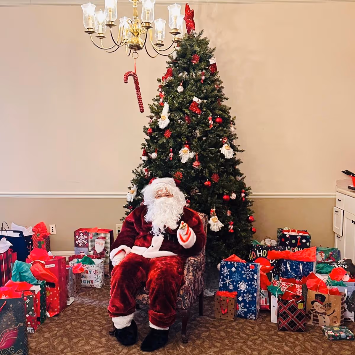 A person dressed as Santa Claus sitting in a chair in front of a decorated Christmas tree surrounded by numerous wrapped gift bags and presents in a room with beige walls and carpeted floor.