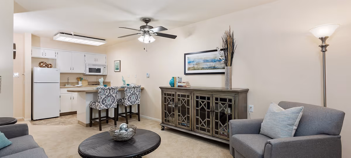 Open living room with armchairs, coffee table, a console cabinet, and an adjacent kitchenette with bar stools and ceiling fan.
