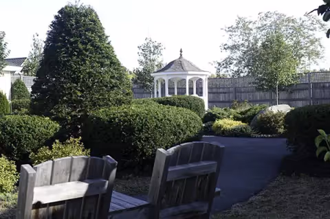 Well-maintained garden with trimmed bushes, two wooden benches in the foreground and a white gazebo in the background.