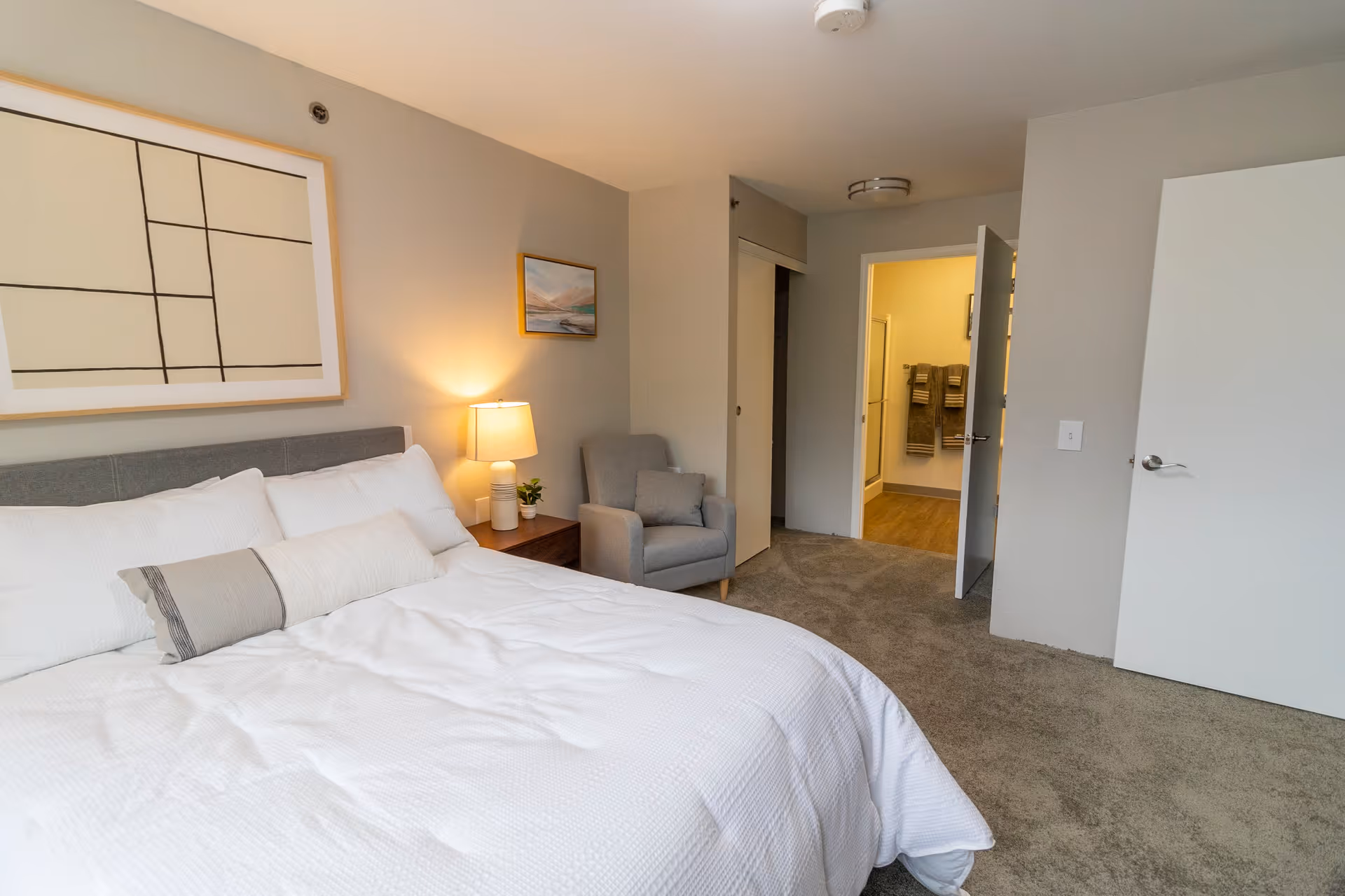 A bedroom with a neatly made bed featuring white bedding and pillows. Next to the bed is a wooden nightstand with a lamp and a small plant. A gray armchair is positioned against the wall near an open door leading to a bathroom with visible towels hanging. The walls are painted light gray and decorated with two framed artworks.