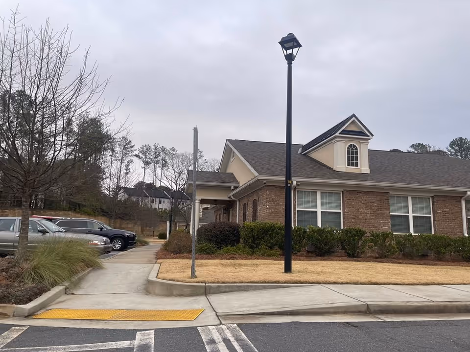 Front exterior of a single-story brick building with windows, a lamp post, parking spaces, and bare trees under a cloudy sky.