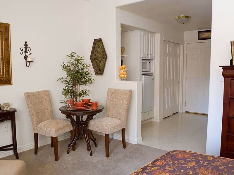 A cozy interior corner of a senior living facility featuring a small round wooden table set with red dishes and two beige upholstered chairs. A green potted plant is placed on the table. The background shows a partial view of a kitchen area with white cabinets, a microwave, and a refrigerator. The floor transitions from carpet in the seating area to tile in the kitchen and hallway. The walls are white with decorative wall hangings and a framed picture.