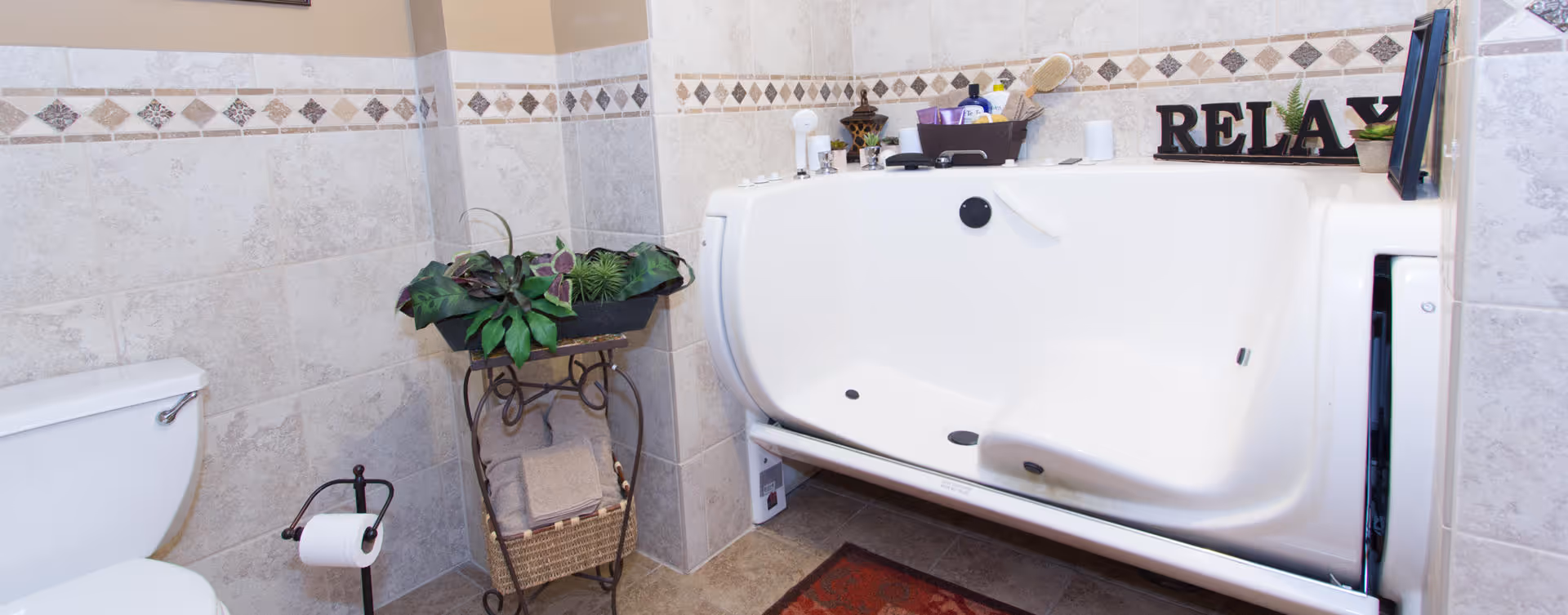 Tiled bathroom featuring a walk-in bathtub, toilet, decorative plant stand and 'RELAX' letters on the tub ledge.