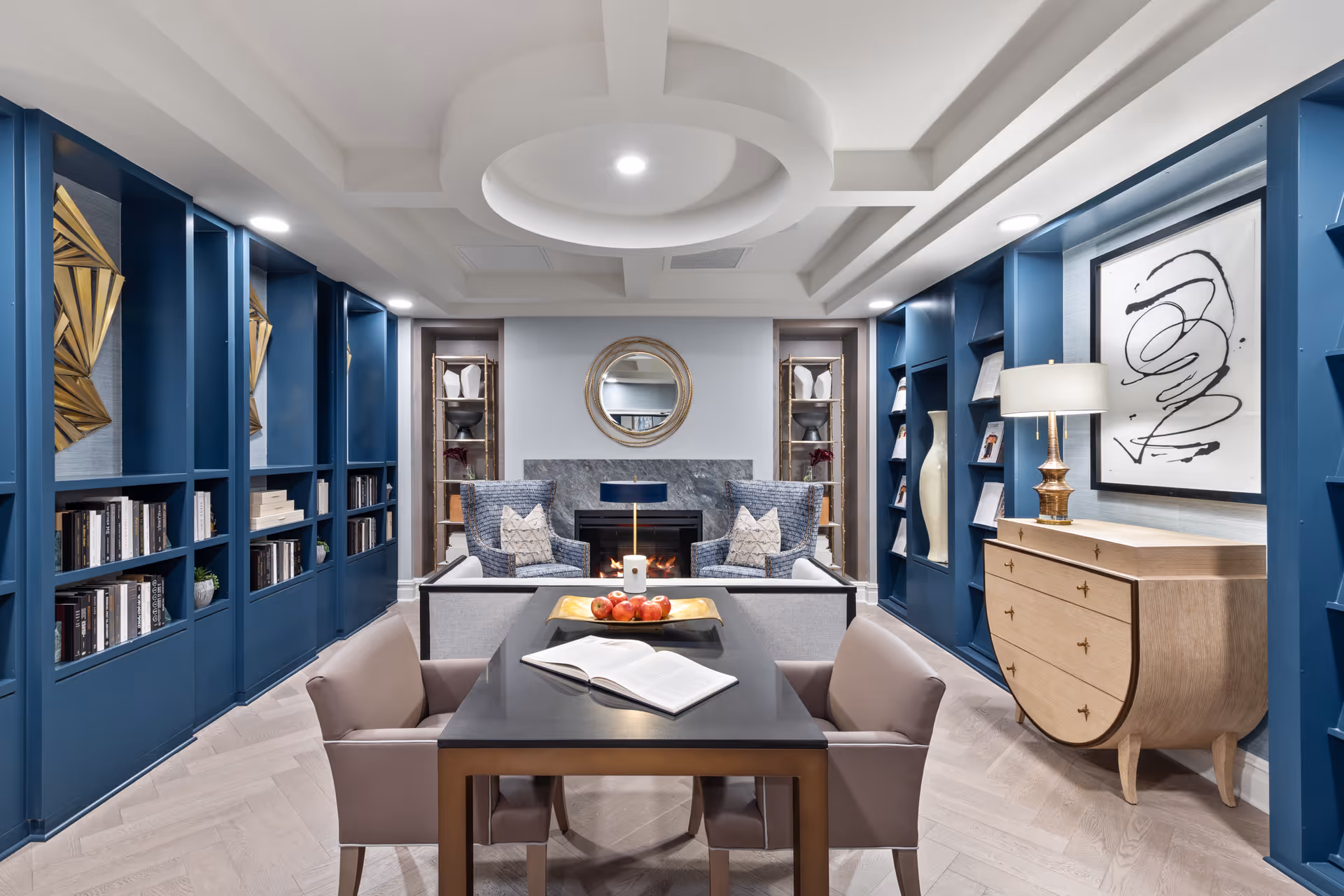 A modern and elegant living room with blue built-in bookshelves on both sides, a black table with four beige chairs in the center, an open book and a tray with apples on the table. At the back, there are two blue armchairs with patterned pillows in front of a fireplace with a round mirror above it. A wooden dresser with a lamp and abstract artwork is on the right side.