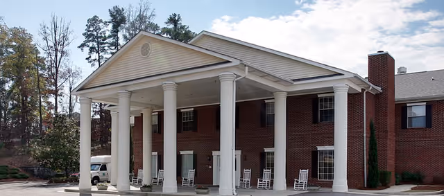 Front exterior view of a two-story brick building with white columns supporting a large covered entrance. Several white rocking chairs are placed on the porch. Trees and a partly cloudy sky are visible in the background.