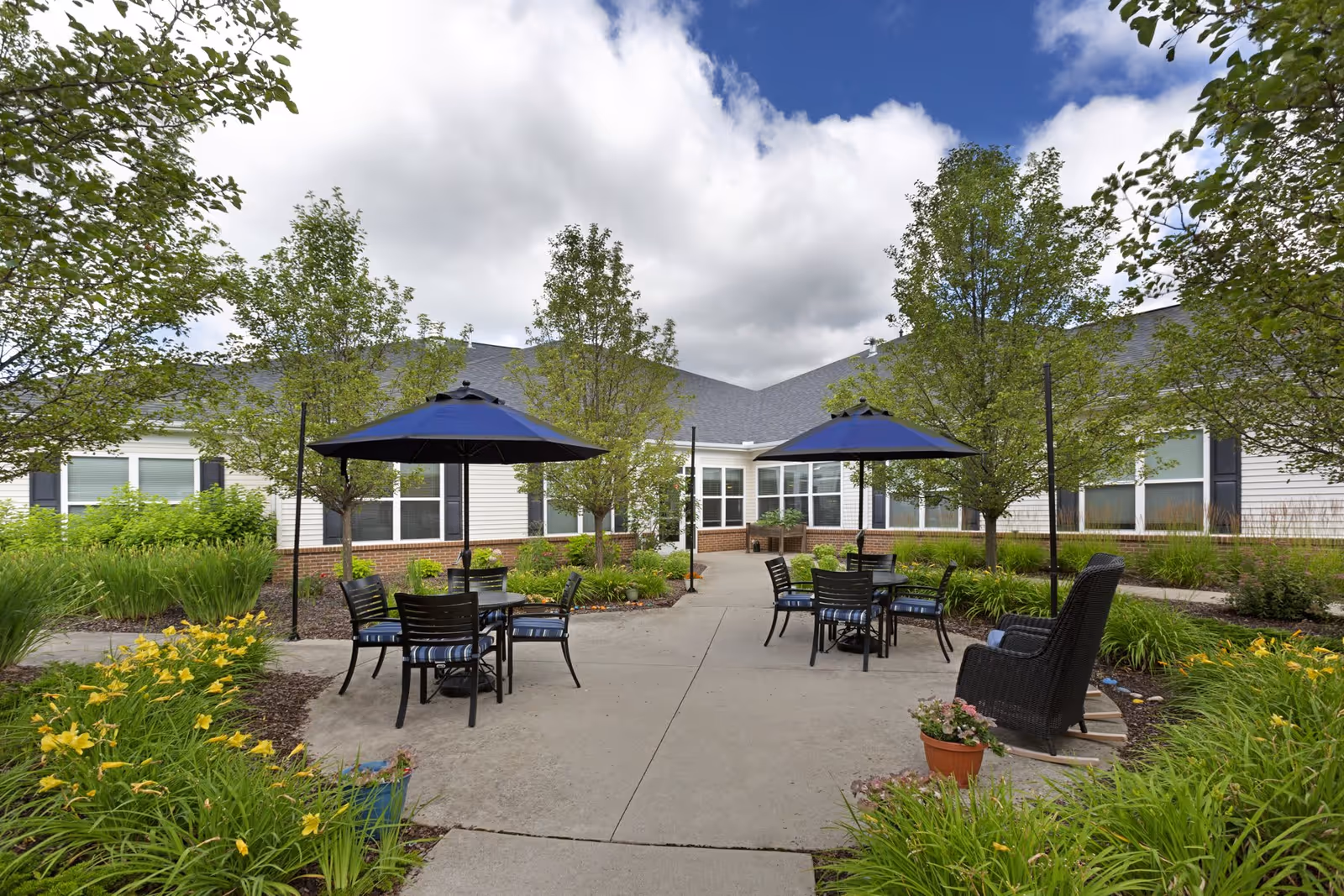 Outdoor courtyard area at Olivia's Assisted Living with concrete pathways, several tables with blue umbrellas, black chairs with blue cushions, surrounded by green trees, bushes, and yellow flowers under a cloudy sky.