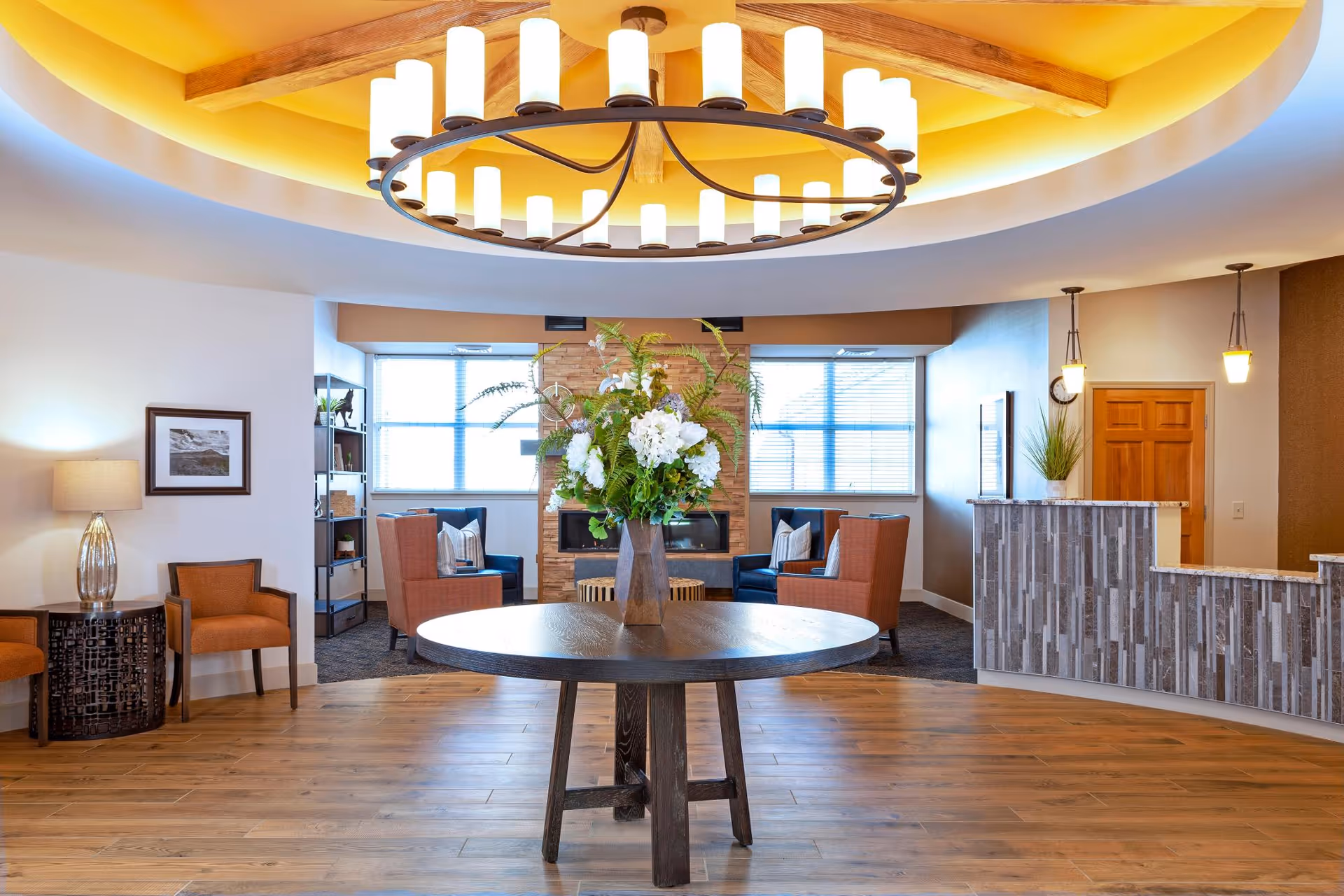 A spacious and well-lit common area featuring a round wooden table with a large floral arrangement in the center. Surrounding the table are comfortable armchairs arranged near a modern fireplace with a brick surround. The room has large windows allowing natural light, a wooden floor, a decorative ceiling with a circular light fixture, and a reception desk on the right side.