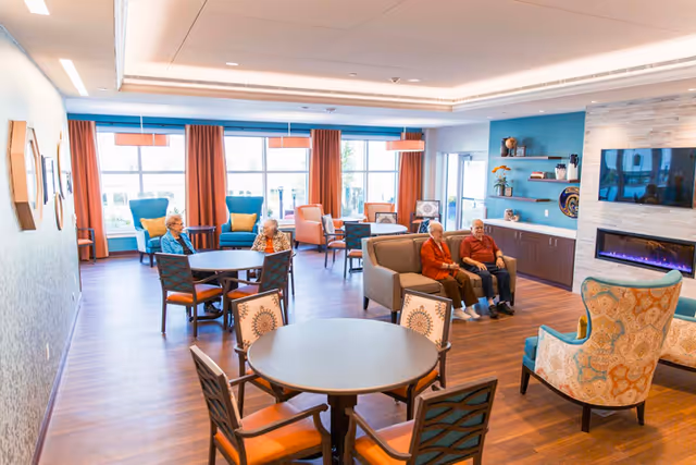 A bright and spacious senior living common area with several round tables and chairs, a modern fireplace, a wall-mounted TV, and large windows with orange curtains. Four elderly people are seated, two on a sofa and two at a table, engaging in conversation.