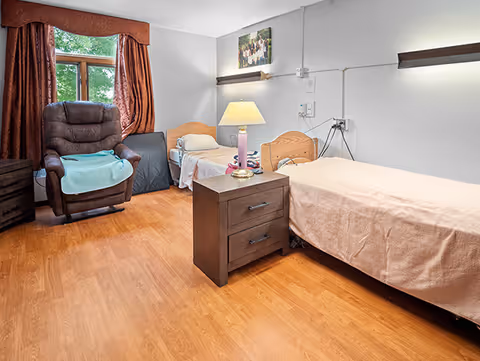 A senior living facility bedroom with two single beds, each with light-colored bedding. Between the beds is a dark wooden nightstand with a lamp on it. There is a comfortable recliner chair with a blue blanket draped over the armrest near a window with brown curtains. The room has wooden flooring and light-colored walls with a photo hanging above one bed.