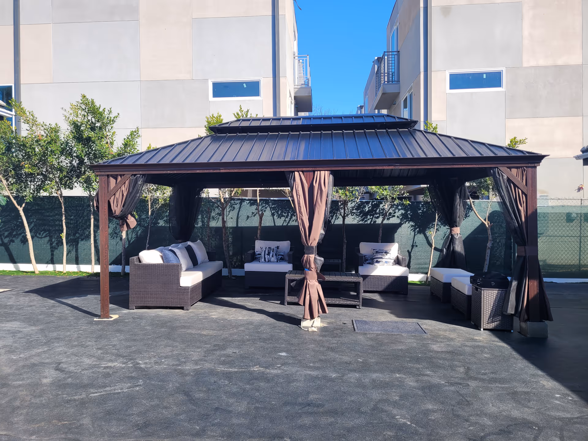 Covered outdoor gazebo with wicker sofas and chairs arranged on a paved courtyard in front of a building.