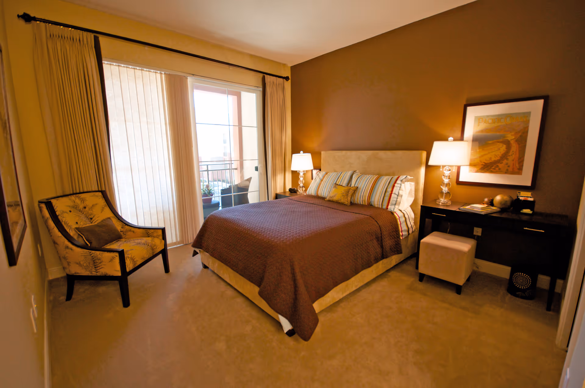 A cozy bedroom with a bed covered in a brown quilt and striped pillows, two bedside tables each with a lamp, a framed picture on the wall, a cushioned chair with a floral pattern, and sliding glass doors leading to a balcony.