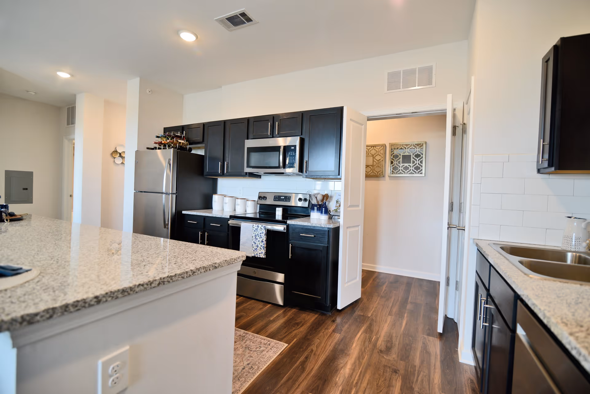 Modern kitchen with dark wood cabinets, stainless steel refrigerator, stove, and microwave. Granite countertops with a double sink on the right and a large island countertop on the left. Hardwood flooring and white walls with two decorative framed artworks visible through an open door.