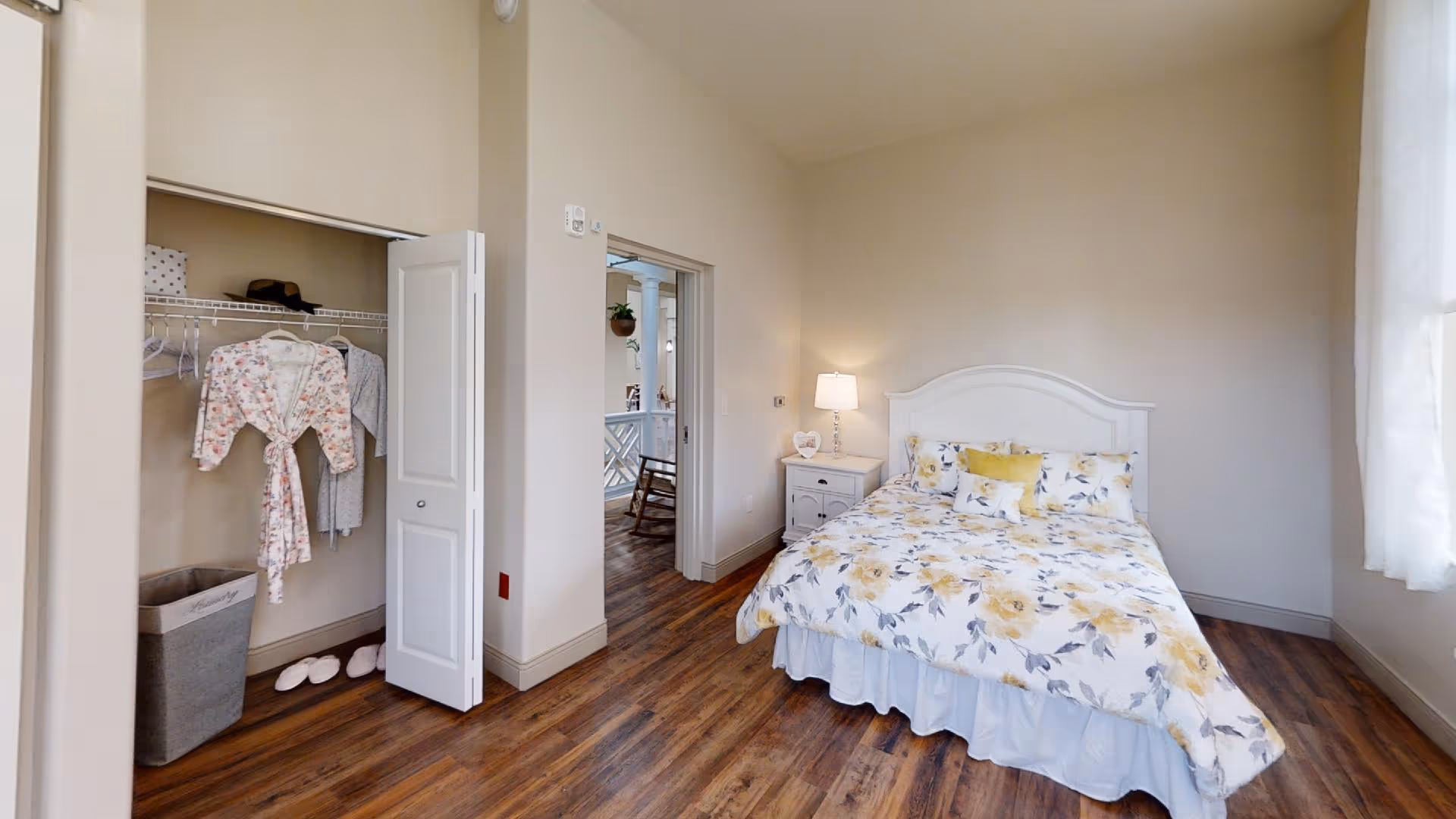 A cozy bedroom with a bed covered in a floral yellow and white comforter, a white nightstand with a lamp, an open closet with hanging robes and slippers, and wooden flooring. A window with sheer curtains lets in natural light.