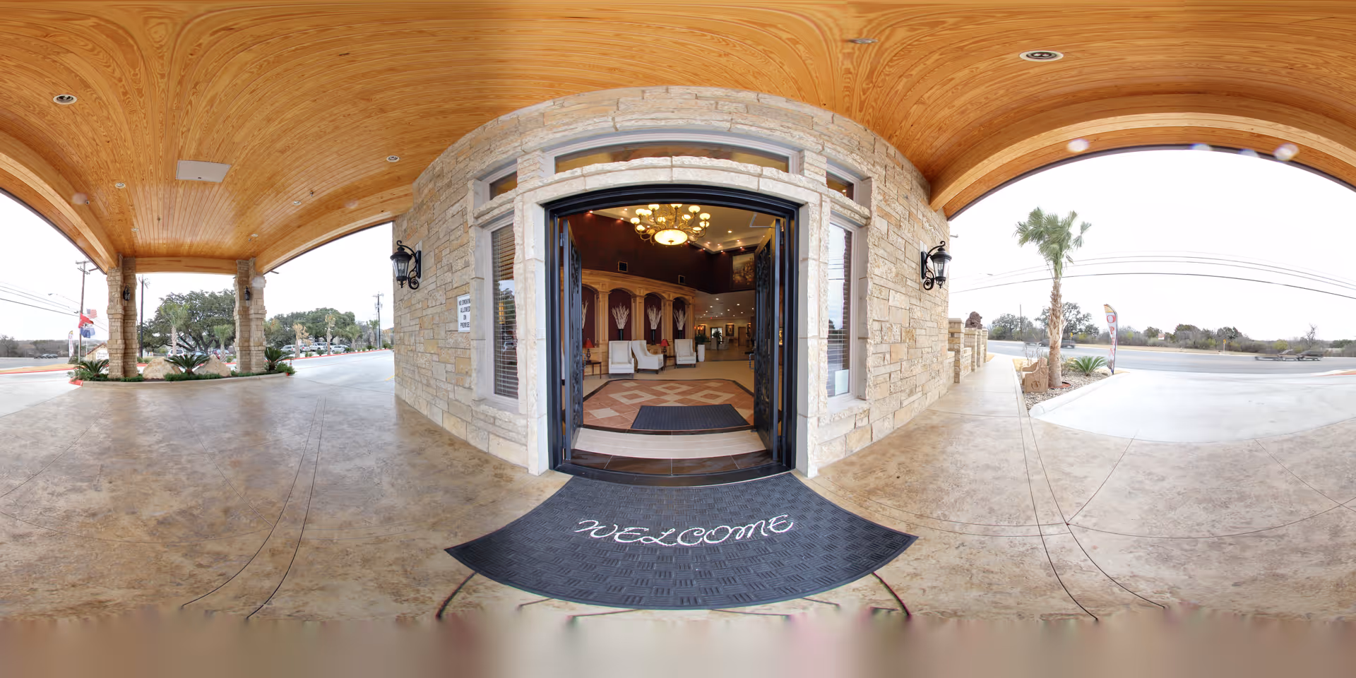 Covered entrance area of a building with a stone facade and wooden ceiling. Double glass doors are open, revealing a warmly lit interior with chairs and a chandelier. A black welcome mat is placed at the entrance. Outside, there are palm trees and a paved driveway.