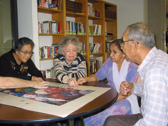 Four elderly individuals sitting around a round table working together on assembling a large jigsaw puzzle in a room with bookshelves filled with books in the background.