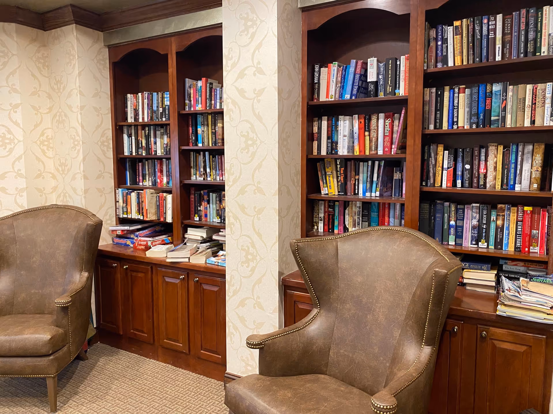A cozy reading area with two brown leather armchairs facing built-in wooden bookshelves filled with books and some board games on the lower shelves. The walls have a beige patterned wallpaper and the floor is carpeted.