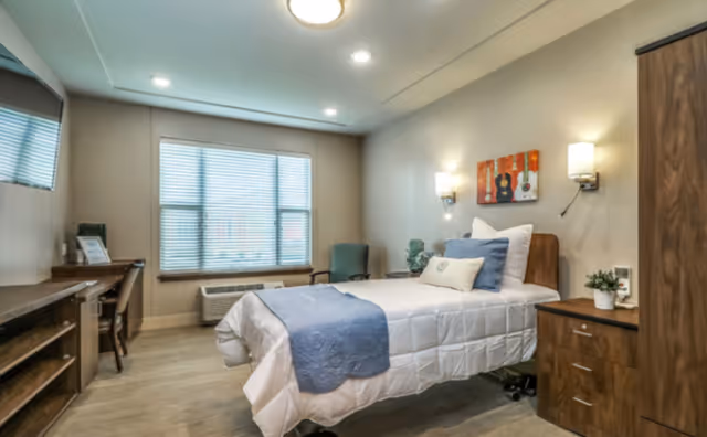 A well-lit bedroom in a senior living facility featuring a single bed with white bedding and a blue throw, a wooden nightstand with a small plant, a wooden wardrobe, a desk with a chair, a green armchair near the window, and wall-mounted lights above the bed. The room has a large window with blinds and neutral-colored walls.