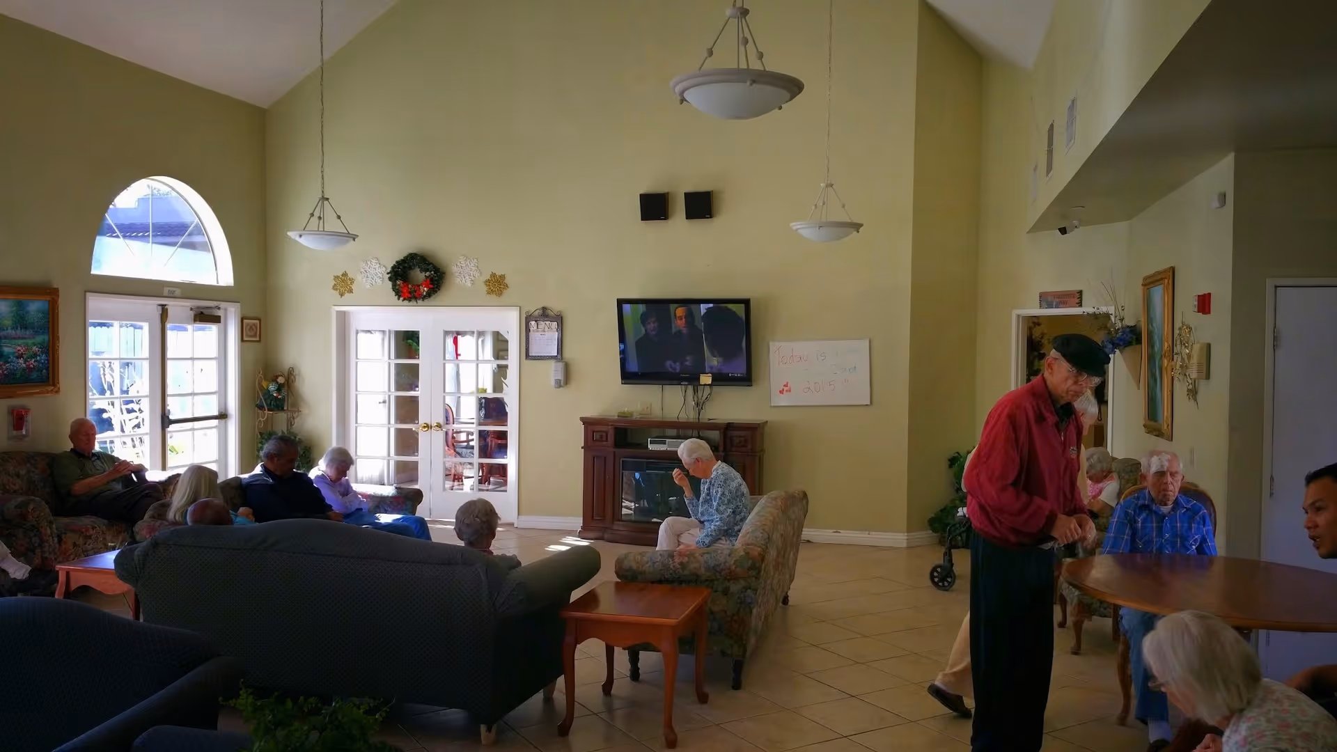 A common area in a senior living facility with several elderly people sitting on couches and chairs, watching a television mounted on the wall above a fireplace. The room has high ceilings, pendant lights, and large windows letting in natural light. There are decorations on the walls including a wreath and framed pictures.