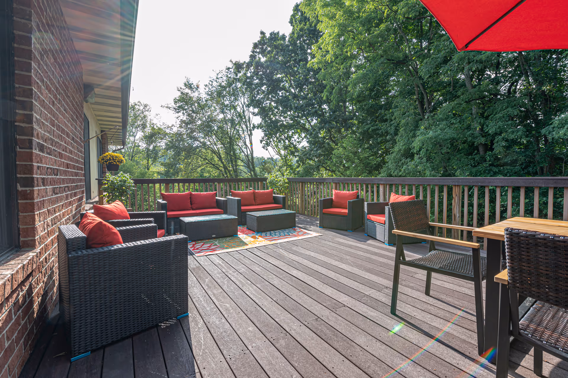Outdoor wooden deck with dark wicker patio furniture featuring red cushions, a colorful rug, and surrounded by green trees under a sunny sky.