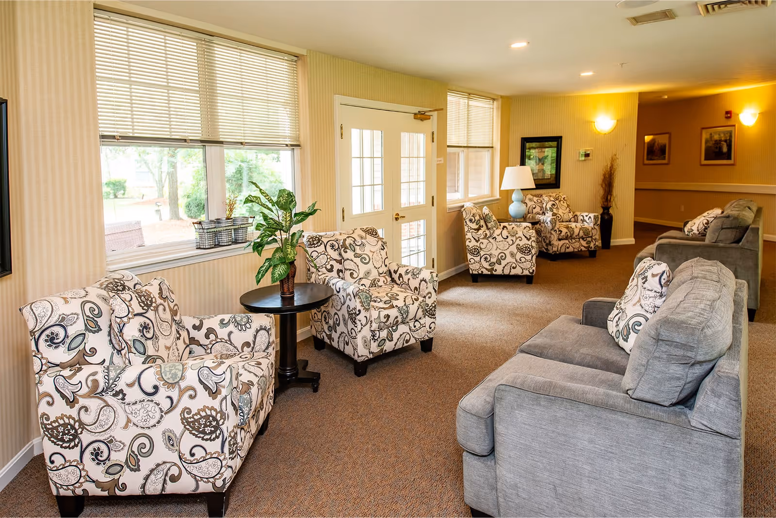 A cozy living room area in a senior living facility with patterned armchairs and gray sofas arranged around small tables. The room has beige walls, carpeted floors, large windows with blinds, and soft lighting from wall sconces and a table lamp. There are framed pictures on the walls and a potted plant on a table near the window.